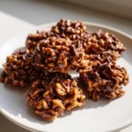 A stack of rich, chocolate-covered haystack cookies made with crispy rice cereal, glistening under sunlight.