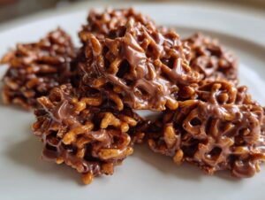 A close-up of several glossy, chocolate-coated haystack cookies piled on a white plate.