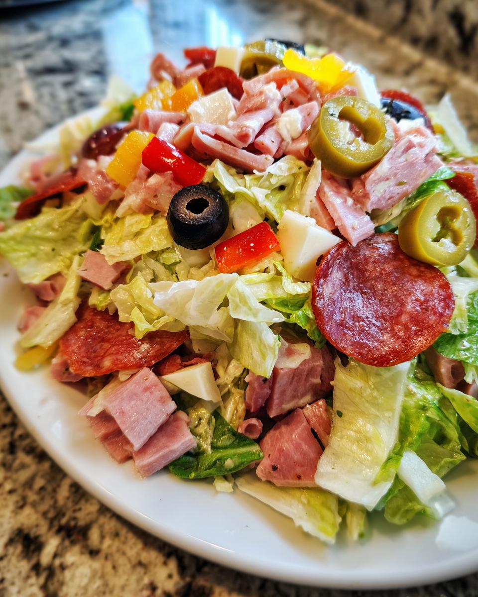 A close-up view of a hearty grinder salad featuring chopped lettuce, salami, ham, cheese cubes, olives, and peppers.