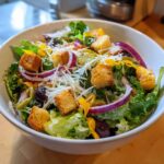 A vibrant bowl featuring a copycat Olive Garden salad with crisp lettuce, red onion rings, black olives, croutons, and shredded Parmesan.