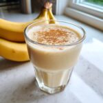 A creamy banana smoothie in a glass, dusted with cinnamon, with fresh bananas in the background.