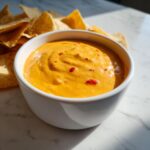 A white bowl filled with bright orange cottage cheese queso, topped with red pepper flakes, next to tortilla chips.
