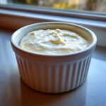 A small white ramekin filled with thick, creamy homemade garlic sauce, sitting on a windowsill.