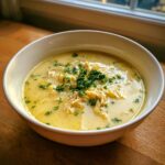 A creamy, yellow bowl of lemon chicken soup filled with shredded chicken and topped with fresh parsley.