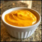 Close-up of bright orange, creamy nacho cheese sauce served in a small white fluted ramekin.