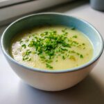 A close-up of creamy potato leek soup in a light blue rimmed bowl, garnished generously with fresh chopped chives.