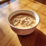 A white bowl filled with creamy white chicken chili, topped with shredded white chicken and beans, sitting on a sunlit wooden table.