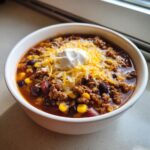 A white bowl filled with rich crockpot cowboy soup, topped with shredded cheese and a dollop of sour cream.