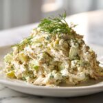 A generous mound of creamy dill pickle chicken salad garnished with fresh dill sprigs on a white plate.