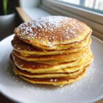 A tall stack of golden brown coconut flour pancakes dusted heavily with powdered sugar on a white plate.