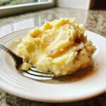 A serving of creamy, fluffy garlic mashed potatoes topped with melted butter and roasted garlic pieces on a white plate.