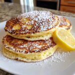 A stack of three fluffy ricotta pancakes dusted heavily with powdered sugar, served with a lemon wedge.