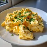 A close-up of incredibly fluffy scrambled eggs, perfectly cooked and garnished with chopped chives and black pepper.