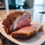 A thick slice of medium-rare prime rib roast with a dark, seasoned crust resting next to the main roast on a white plate.