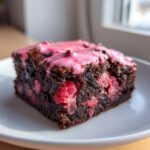 A close-up of a fudgy strawberry brownie slice topped with bright pink glaze, showing chunks of strawberry inside.
