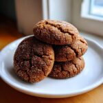 A stack of four chewy, dark brown ginger molasses cookies coated in sparkling sugar crystals on a white plate.