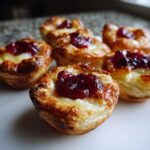 Close-up of several golden baked brie bites made with puff pastry, melted brie, and a dollop of red jam.