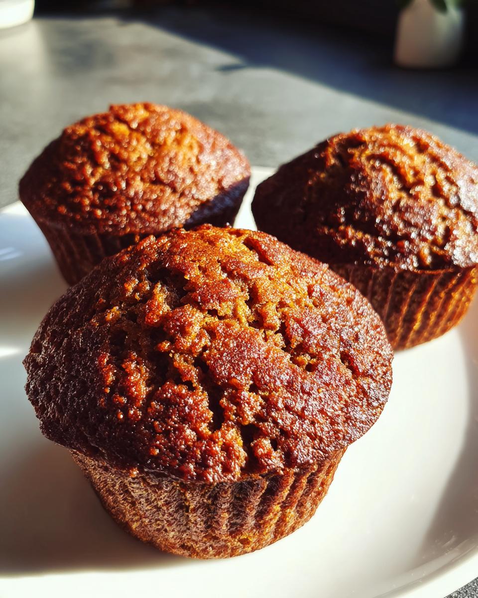 Close-up of three freshly baked, dark golden brown bran muffins sitting on a white plate.