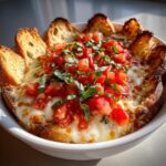 A close-up of a hot, baked bruschetta dip topped with melted cheese, fresh tomatoes, and basil, served with toasted bread pieces.