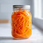 A clear glass mason jar filled tightly with bright orange, julienned pickled carrots, sealed with a silver lid.