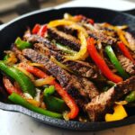 Close-up of juicy steak fajitas with grilled red, green, and yellow peppers and onions in a black skillet.