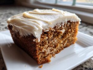 Close-up of a moist slice of applesauce cake topped with thick, creamy frosting, sitting on a white plate.