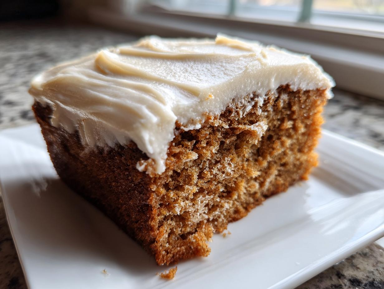 Close-up of a moist slice of applesauce cake topped with thick, creamy frosting, sitting on a white plate.