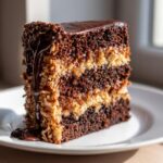 A tall, moist slice of german chocolate cake featuring dark layers and rich coconut-pecan frosting, topped with chocolate ganache.