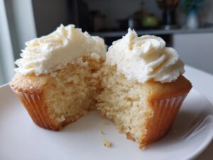 A close-up of a moist vanilla cupcake cut in half, revealing the fluffy interior and topped with piped white frosting.