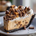 Close-up of a rich slice of no bake butterfinger pie on a white plate, topped with whipped cream and candy pieces.