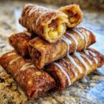 A stack of golden brown, fried peach cobbler egg rolls coated in cinnamon sugar and drizzled with white icing.