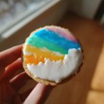 A hand holding a round sugar cookie decorated with bright, layered rainbow colors and white icing clouds, showcasing the sugar cookie icing recipe.
