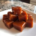 Close-up of glossy, chewy caramel squares piled on a white plate, showcasing the rich caramel color.