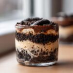 Close-up of a single serving dirt cake layered with cream and crushed chocolate cookies in a glass cup.