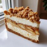 A tall slice of apple pie cheesecake featuring layers of graham crust, creamy cheesecake, spiced apples, and a crumbly topping.