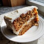 A close-up slice of moist applesauce cake layered with thick white frosting, sitting on a white plate.