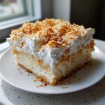 A moist slice of coconut poke cake topped with whipped cream and toasted coconut flakes.