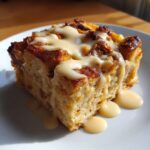 A close-up of a rich slice of bread pudding topped with a thick vanilla cream sauce.