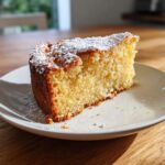 A close-up shot of a moist slice of olive oil cake dusted with powdered sugar, sitting on a white plate.
