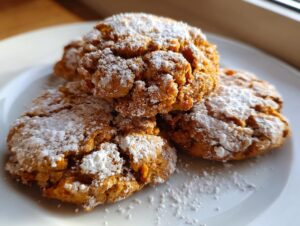 Close-up of three soft persimmon cookies piled on a white plate, heavily dusted with powdered sugar.