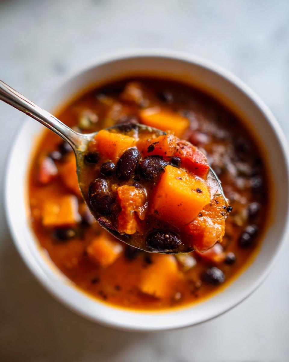 A spoonful lifted from a bowl of rich butternut squash chili, showing chunks of orange squash and black beans.