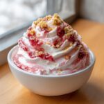 Close-up of a bowl of strawberry cheesecake dip, swirled high with whipped topping, strawberry sauce, and graham cracker crumbs.