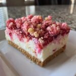 A close-up of a perfect slice of strawberry crunch cheesecake showing the graham cracker crust, creamy filling, and pink crumble topping.