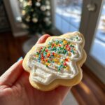 A hand holding a tree-shaped sugar cookie topped with bright white cookie icing and colorful nonpareil sprinkles.
