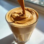 Close-up of a thick, creamy peanut butter banana smoothie being poured into a glass, creating swirls on top.