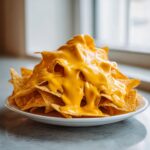 A close-up of tortilla chips completely covered in thick, bright yellow nacho cheese sauce, ready to eat.