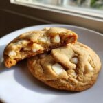 Two freshly baked white chocolate macadamia nut cookies on a white plate, one broken open showing a gooey center.