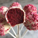Close-up of red velvet cake pops dipped in white chocolate and covered in pink sprinkles, one is bitten open showing the interior.