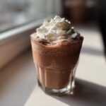 A glass of rich, blended frozen hot chocolate topped with a generous swirl of whipped cream, sitting in sunlight.