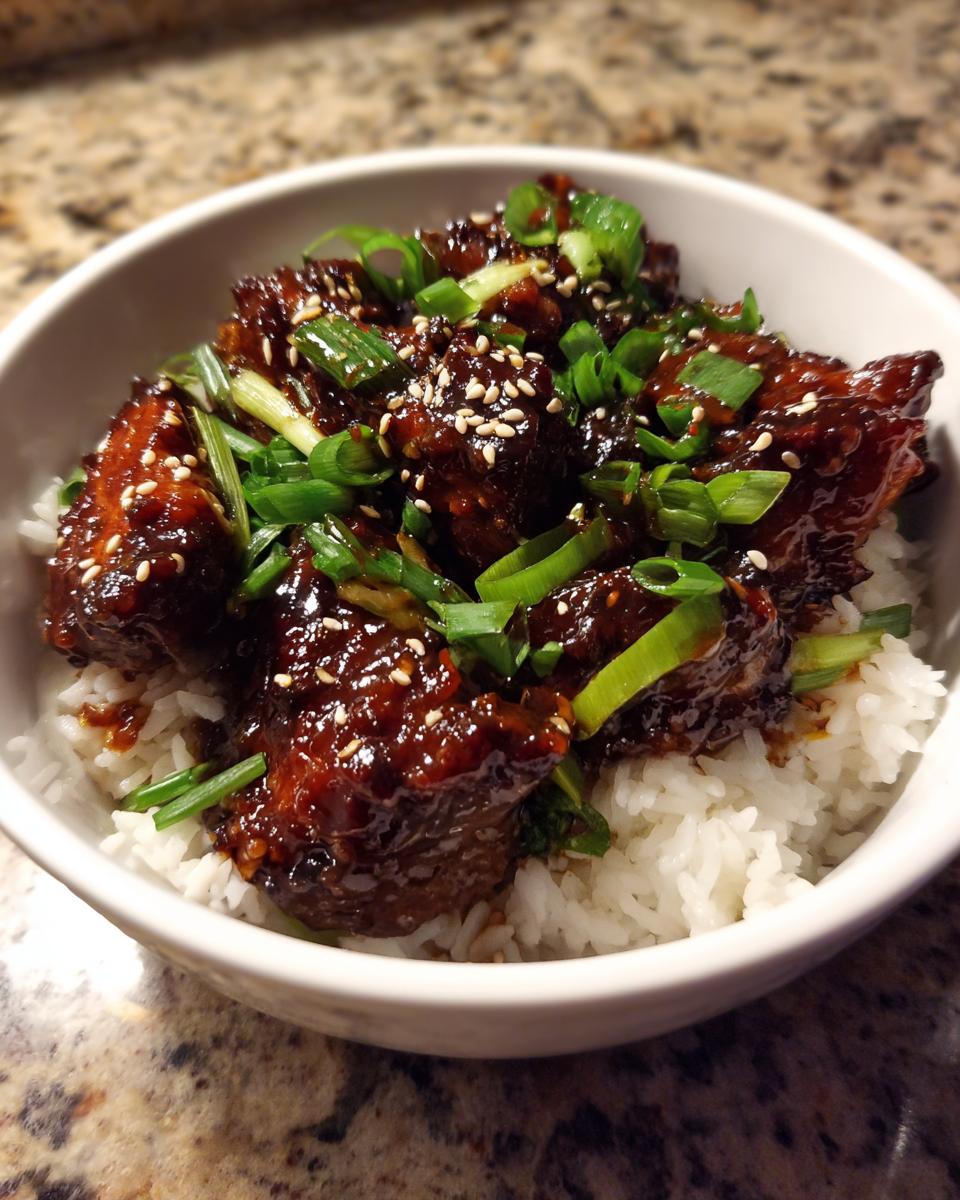 Close-up of a white bowl filled with white rice topped with dark, glossy glazed chicken pieces, green onions, and sesame seeds, making a delicious chicken rice bowl.